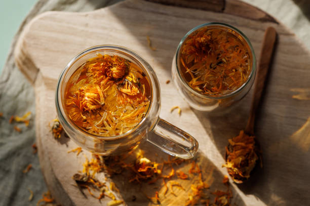 Calendula herbal tea in two glass cups on wooden tray Istanbul, Turkey-September 22, 2022: Calendula herbal tea in two glass cups on wooden tray. There are dried calendula flowers on the tea. There are calendulas scattered around. Double wall glass. Shot with Canon EOS R5. pot marigold stock pictures, royalty-free photos & images