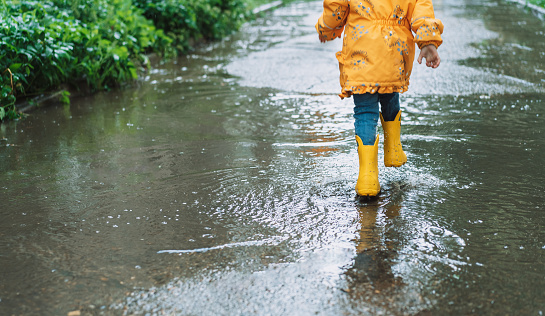 Girl in yellow jacket and rubber boots runs through the puddles.