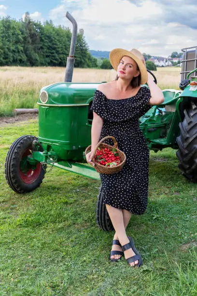 Young woman with a basket of cherries stands near a tractor in the countryside