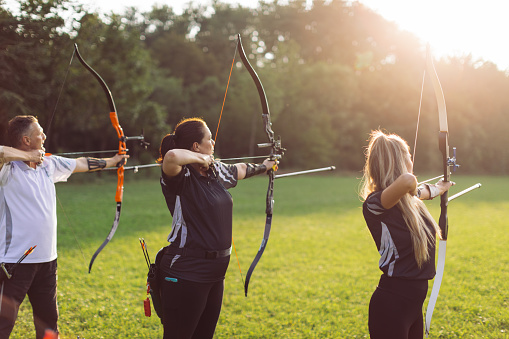 Women and man practicing archery training with recurve bow on open field before sunset.