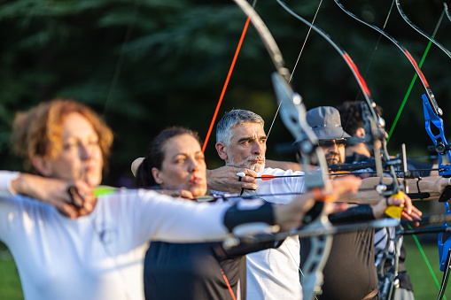 Women and men practicing archery training with recurve bow on open field before sunset.