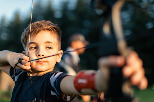 Boy holding bow and arrow and aiming. He is practicing archery outdoors and he is very concentrated
