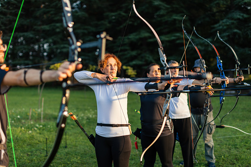 Women and men practicing archery training with recurve bow on open field before sunset.