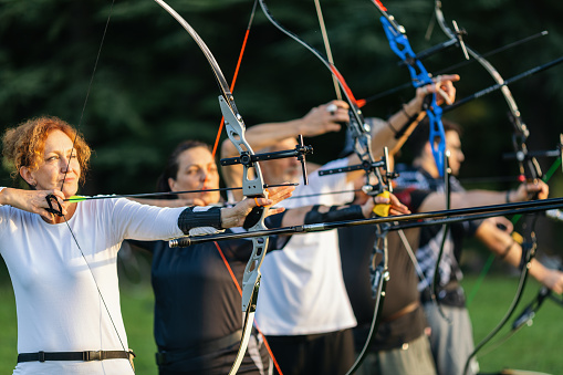 Women and men practicing archery training with recurve bow on open field before sunset.