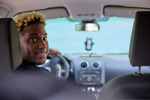 Friendly young African man in casual clothes, looking at the camera over his shoulder and smiling while driving a car. Are you ready to go.