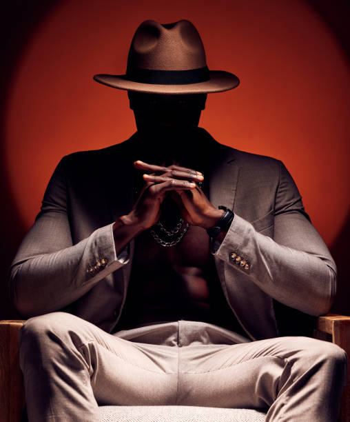 hombre elegante, moderno y moderno en silueta sentado en una habitación oscura con un fondo de estudio rojo. jefe de la mafia masculino vanguardista, misterioso y curioso sentado con un traje elegante, sombrero y joyas caras - fugitive fotografías e imágenes de stock