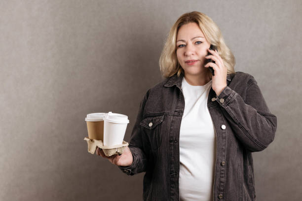 Attractive blonde plus-size with two glasses of coffee talk on the phone on a gray background Attractive blonde plus-size with two glasses of coffee talk on the phone on a gray background. plus size smart casual dresses stock pictures, royalty-free photos & images