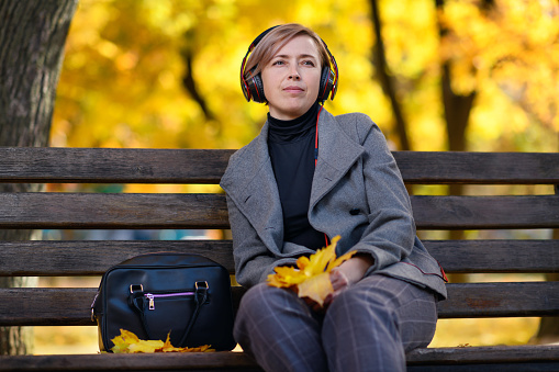 beautiful woman portrait, she is sitting on a bench in the autumn park and listens to music with headphones, trees with yellow leaves, bright sunny day