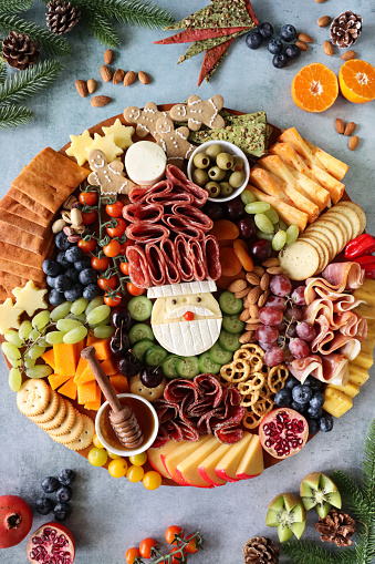 Stock photo showing close-up, elevated view of Christmas design charcuterie board covered with prepared sliced and chopped ingredients including a Brie Father Christmas, salami, cucumber, gingerbread men, star-shaped cheddar cheese, vine tomatoes, red and white grapes, cherries, almonds, apricots, blueberries, pretzels and a ramekins of green olives and honey.