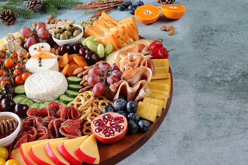 Stock photo showing close-up, elevated view of Christmas design charcuterie board covered with prepared sliced and chopped ingredients including a Brie snowman, Edam, chilli cheddar cheese, gingerbread men, salami, ham, vine tomatoes, pomegranate, red and white grapes, pretzels, cherries, cucumber, almonds, apricots and ramekins of green olives and honey.