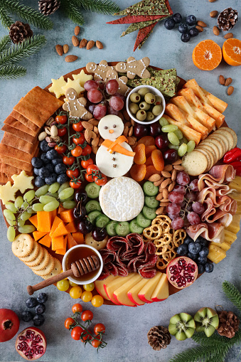 Stock photo showing close-up, elevated view of Christmas design charcuterie board covered with prepared sliced and chopped ingredients including a Brie snowman, Edam, chilli cheddar cheese, gingerbread men, salami, ham, vine tomatoes, pomegranate, red and white grapes, pretzels, cherries, cucumber, almonds, apricots and ramekins of green olives and honey.