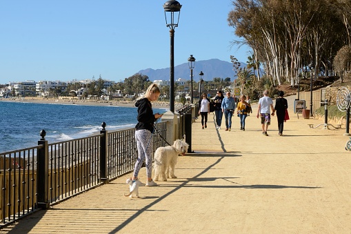Marbella, Spain - December 12, 2021: People walking by the promenade on an autumn sunny day in Marbella, Andalusia, Spain.