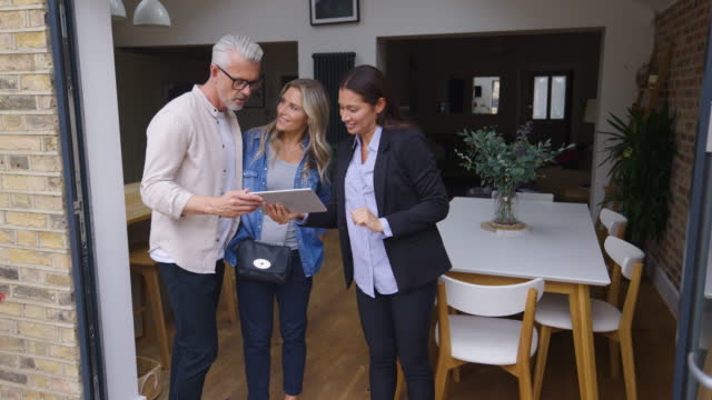 Cheerful real estate agent showing a property to caucasian couple looking at the view and then the tablet to look at the pricing