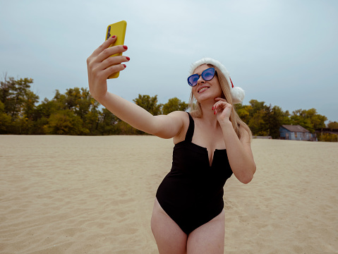 A young woman in a black swimsuit and a New Year's red hat with glasses takes a selfie on a mobile phone in a yellow case while standing on the beach near the forest and far away from the old house. Woman new year. Relax woman. Woman portrait