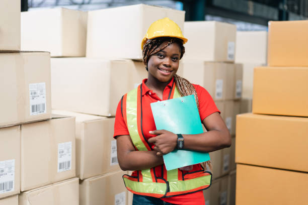 istock Portrait of smiling African female worker holding checklist report document with parcel boxes in the background looking at camera. Warehouse woman working for logistic business 1426293785