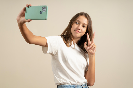 Close-up portrait of a cheerful spanish girl teenager schoolchild pupil taking selfie photo self-picture on smart phone, having video call conversation online isolated in beige background