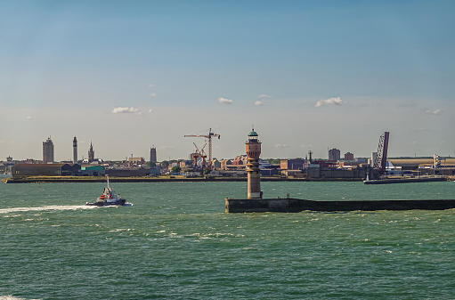 Europe, France, Dunkerque - July 9, 2022: Port scenery. Feu de Saint Pol light tower on its pier at entrance with city skyline on horizon. Belfry towers and Tall Risban light tower. Tugboat on water