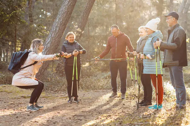 Smiling woman sports trainer in squat position showing technic to people posture of hands, legs for Nordic walking with sticks in forest. Exercises hiking education for senior people. Leisure activity