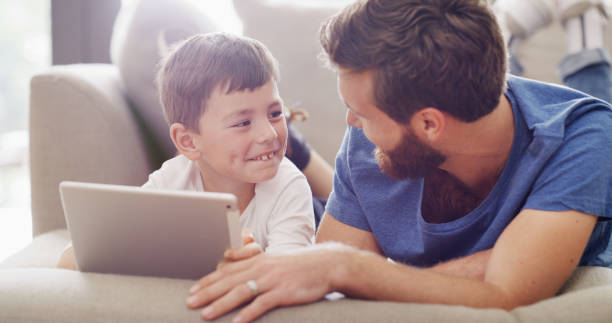 istock Child and father learning and play on a tablet app, while sitting with browsing online in family home living room together. Proud smiling man teaching via a game with his son on a touchscreen 1426303169
