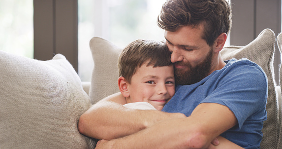 Father, son and hug while bonding on sofa in a house living room with love, trust and security. Portrait of smile, happy and safety with man and boy in family home support and relax on a lounge couch
