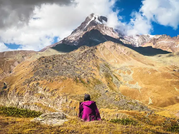 Thoughtful woman sit on grass admire beautiful KAzbek mountain snowy peak in autumn. Hike to gergeti glacier route solo. Travel in caucasus concept