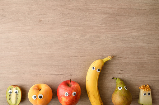 Background of row of funny fruit with eyes on wooden bench as a concept for motivating children to eat. Top view.
