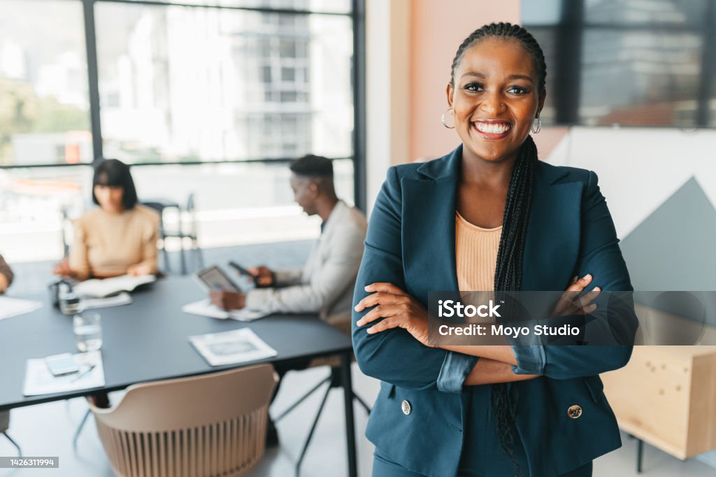 African leader, manager and CEO with a business woman in the office with her team in the background. Portrait of a female boss standing arms crossed at work during a meeting for planning and strategy Leader, manager and CEO with a business woman in the office with her team in the background. Portrait of a female boss standing arms crossed at work during a meeting for planning and strategy Business Person Stock Photo