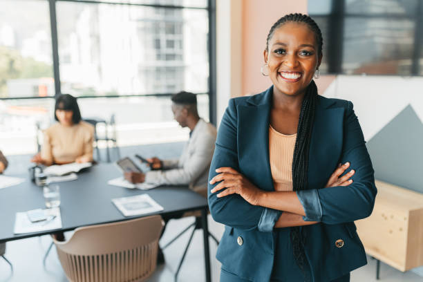 African leader, manager and CEO with a business woman in the office with her team in the background. Portrait of a female boss standing arms crossed at work during a meeting for planning and strategy stock photo