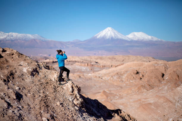 Woman in the Atacama desert looking with binoculars at Licancabur volcano, and at the Kimal mountain. The Atacama Desert is a desert plateau in South America covering a 1,600 km strip of land on the Pacific coast, west of the Andes Mountains.