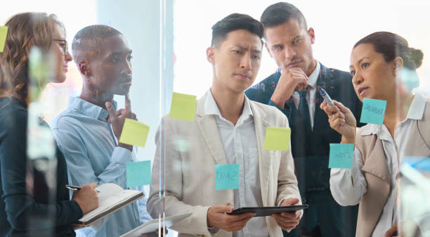 istock Teamwork, strategy and collaboration with planning business  people writing sticky notes on a board or window. Serious thinking and creative problem solving with diverse group on a innovation mission 1426338019