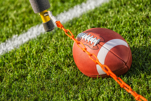 Measuring for a first down next to a college or high school leather American football with a white stripe sitting between the hash marks.\nAmerican and high school use footballs with a white stripe on the top of the ball, whereas the NFL does not have any stripe.