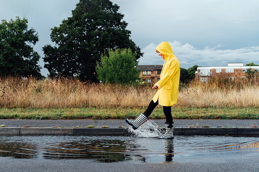 Woman having fun on the street after the rain. Smiling woman wearing rain rubber boots and yellow raincoat walking into puddle with water splash and drops. Fall weather. Selective focus.