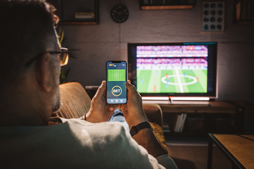 Mature men sitting on sofa at home and watching soccer game.  He is using smart phone for sports betting .