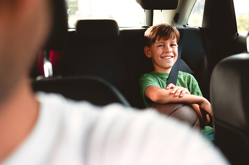 The boy is sitting in the back seat of the car, wearing a seat belt, and holding a basketball