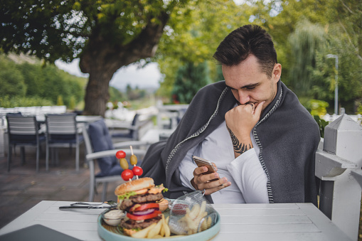 A young man is sitting in the garden of a restaurant and eating a hamburger, enjoying a beautiful autumn day