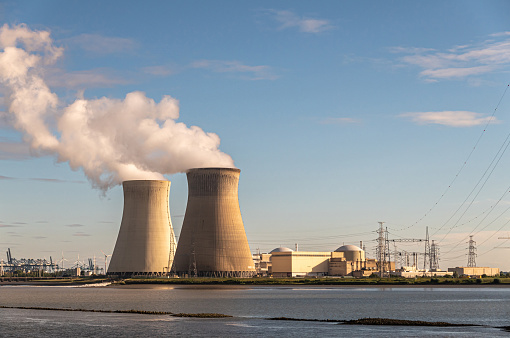 Antwerpen, Flanders, Belgium - July 10, 2022: Sunset on Cooling towers and Doel Nuclear Power plant under blue sky and Scheldt river up front. Container terminal in bottom left corner
