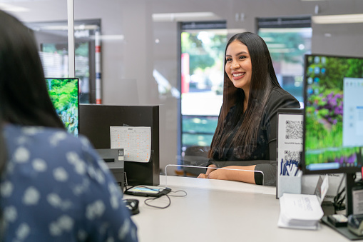 A cheerful and beautiful Hispanic woman smiles while talking to customer service representative at medical office or bank reception desk outfitted with plexiglass sneeze guard.
