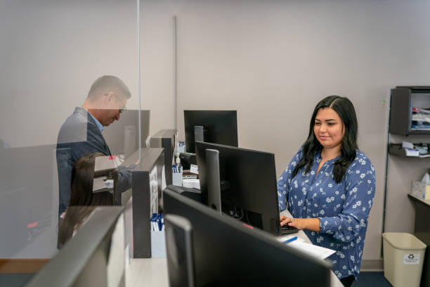 Providing customers with top notch service A beautiful Hispanic woman cheerfully assists a male customer while working at the reception desk of a medical office or bank. bank teller stock pictures, royalty-free photos & images