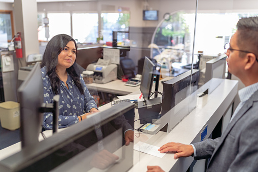A female Hispanic bank teller smiles while helping a male customer at the bank counter.