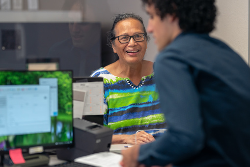 Smiling woman of Pacific Islander descent talks with a helpful customer service representative at the front desk of a medical clinic or bank.