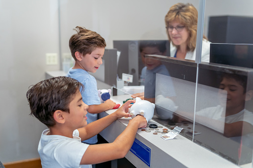 Two elementary age boys count coins from their piggy bank with the assistance of a bank teller when making a deposit at the bank.