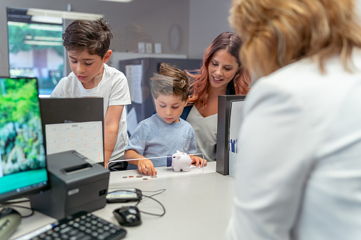 A woman of Latin descent visits the bank with her two young boys and talks with a female bank teller. The children have brought their piggy bank and are planning to open a savings bank account.