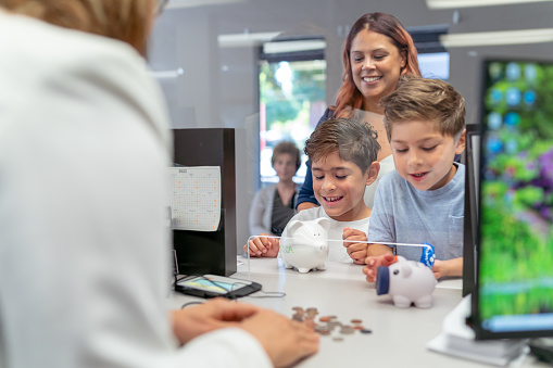 Two elementary age boys of Hispanic descent smile with excitement as a bank teller counts the coins they brought from their piggy banks. The children are learning about money and are opening a bank account with their mom's help.