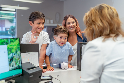 A Hispanic woman and her two young boys smile and greet a female bank teller. The children have brought their piggy bank and plan to open a savings bank account.