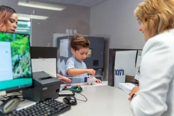 Photo of Cute little boy with piggy bank talking to bank teller