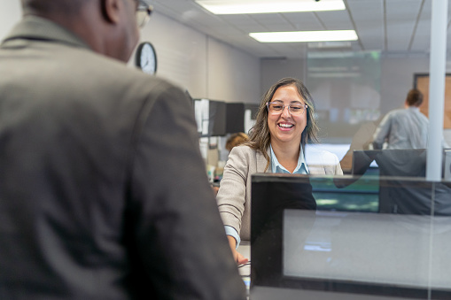 A female customer service representative of Latin descent smiles while greeting an unrecognizable male black customer at the reception desk of a medical clinic or bank.