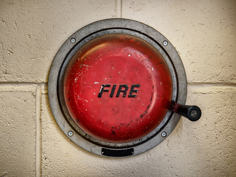 Close up of a  red, vintage Fire alarm, with a black handle, on a cream coloured wall.