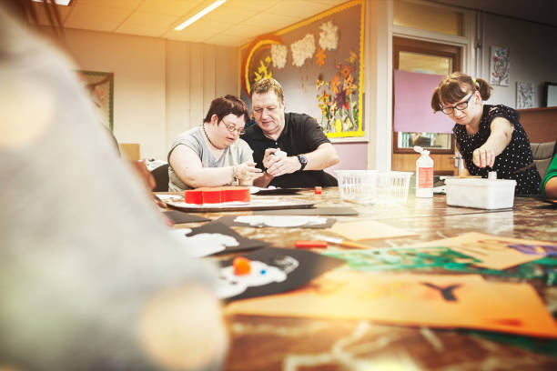 istock Education, development and mental disability, man teaching disabled people. Teacher or volunteer helping men and women with special needs. Learning, support and students in arts and crafts workshop. 1426386754