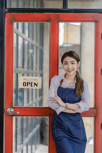 Attractive Asian woman business owner wearing apron and opening red cafe door sign looking at camera, food and beverage business concept.
