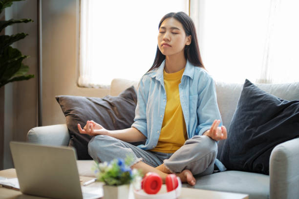 Young asian woman meditating while sitting on couch at home. stock photo